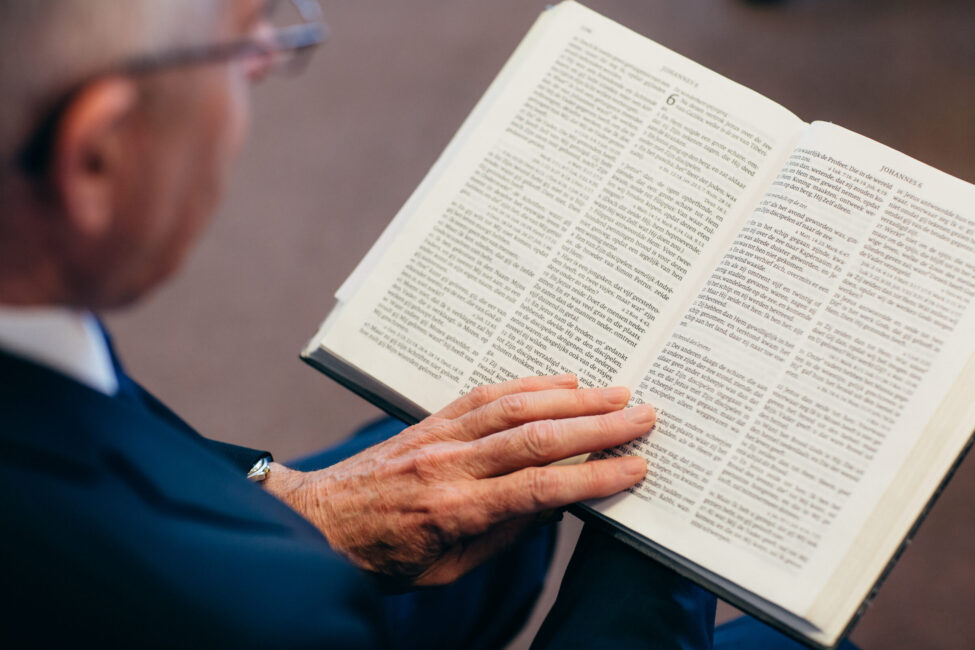 Een oudere man leest aandachtig in een open boek. Hij draagt een donkerblauw pak en zijn handen rusten op de pagina's.