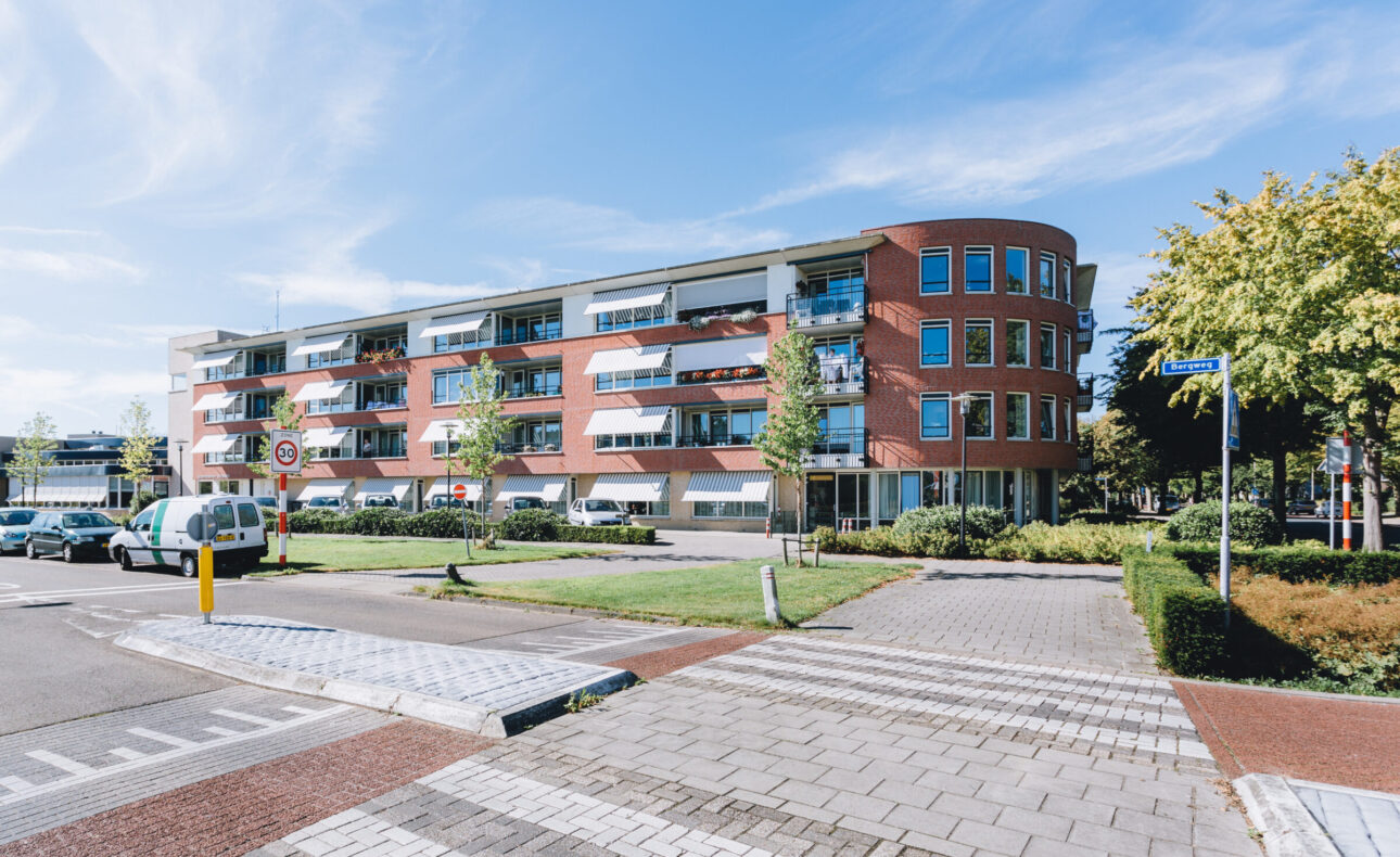 Een modern, rood bakstenen gebouw met meerdere verdiepingen en balkons, omgeven door groen en een blauwe lucht erboven.