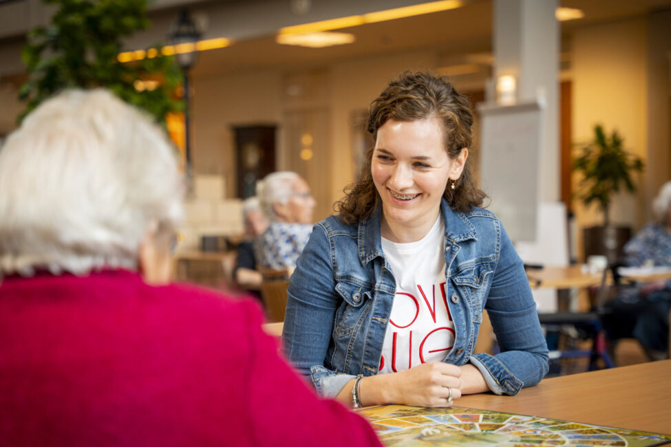Een jonge vrouw glimlacht terwijl ze met een oudere vrouw aan tafel zit in een gemeenschappelijke ruimte van een zorginstelling.