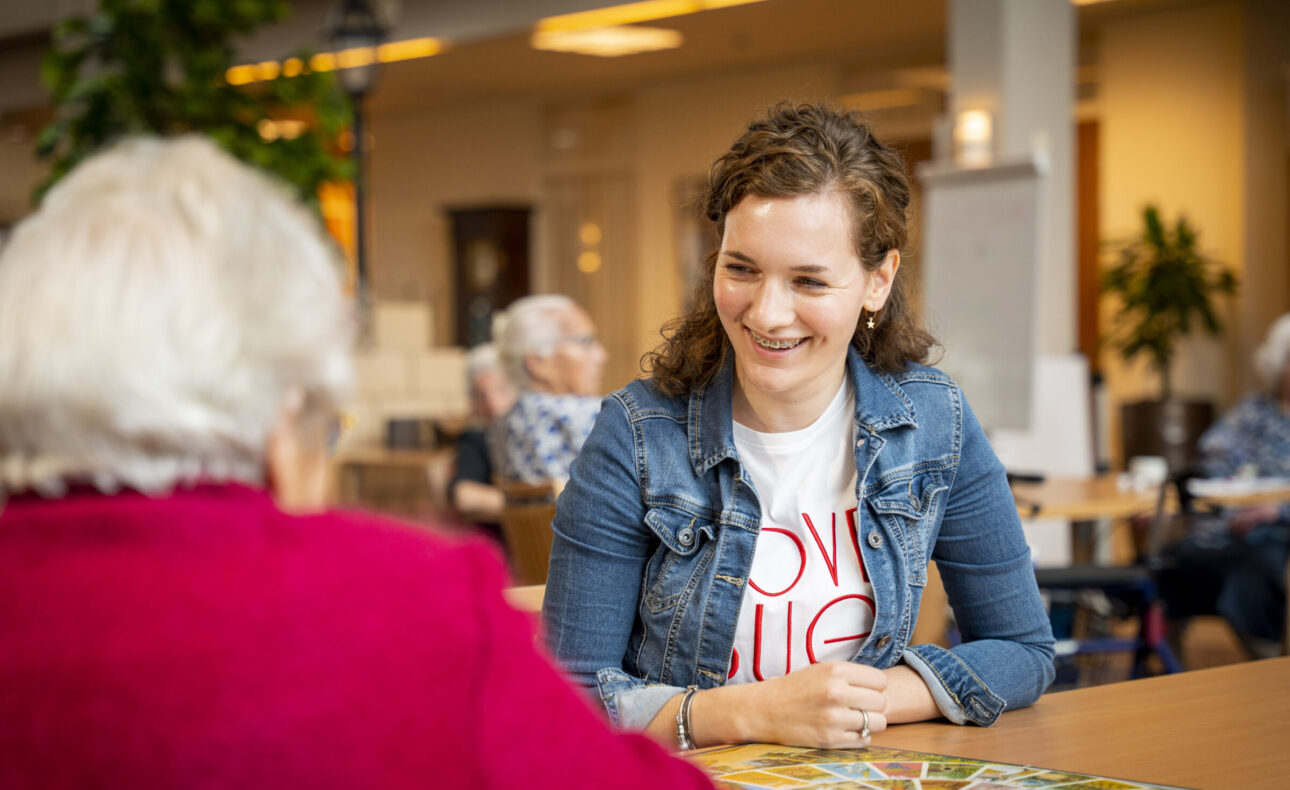 Een jonge vrouw glimlacht terwijl ze met een oudere vrouw aan tafel zit in een gemeenschappelijke ruimte van een zorginstelling.