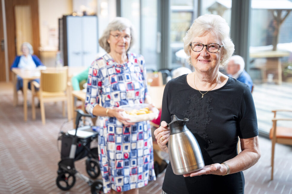 Twee oudere vrouwen in een gemeenschappelijke ruimte. De voorste vrouw houdt een thermoskan vast, de ander een schaal met eten.