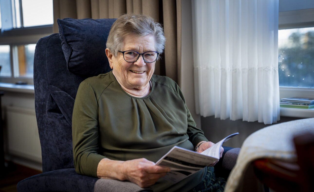 Oudere vrouw met bril zit glimlachend in een stoel bij het raam en leest een tijdschrift in een gezellige kamer.