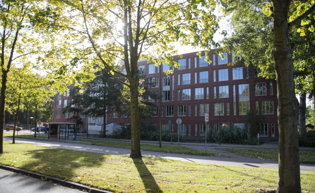 Buitenaanzicht van een modern, rood bakstenen gebouw omgeven door bomen en gras, met een bushalte aan de straatkant.