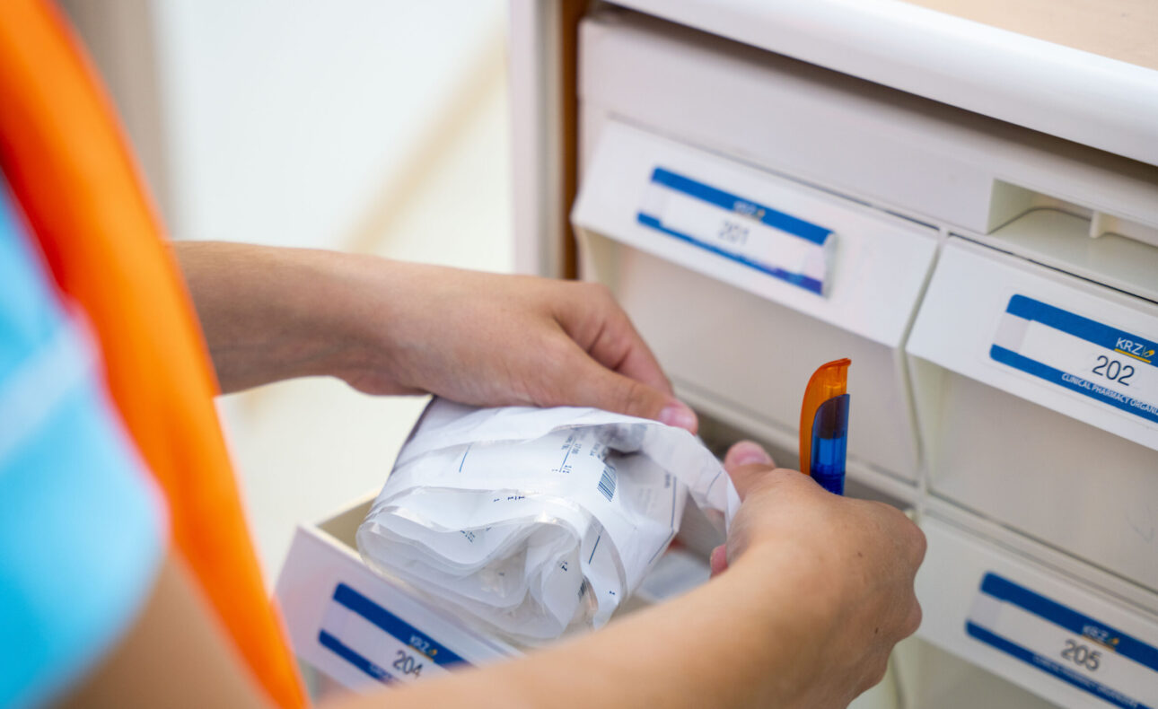 Persoon haalt een pakketje uit een lade met labels in een kast. De persoon draagt een blauw shirt en een oranje hesje.