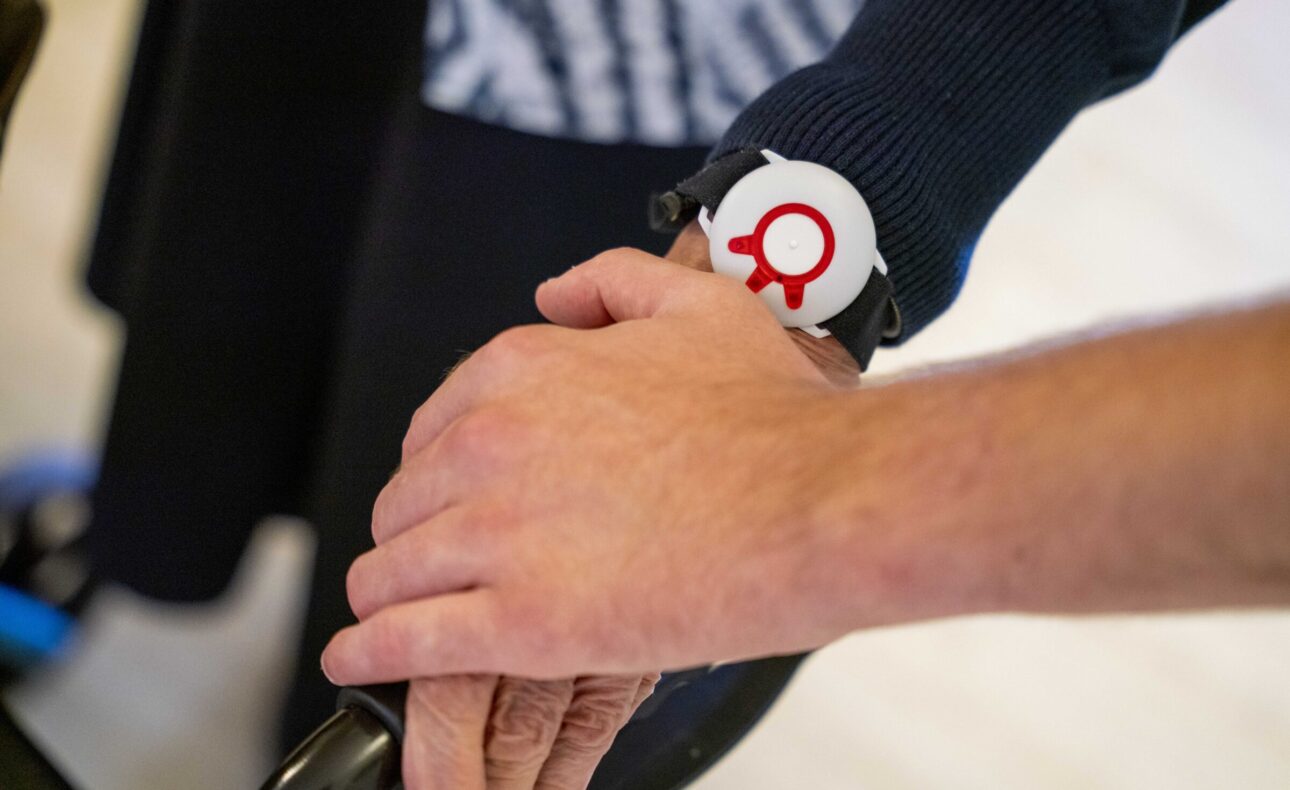 Twee handen houden elkaar vast, waarbij een persoon een zwart polsbandje met een rood-wit symbool draagt.