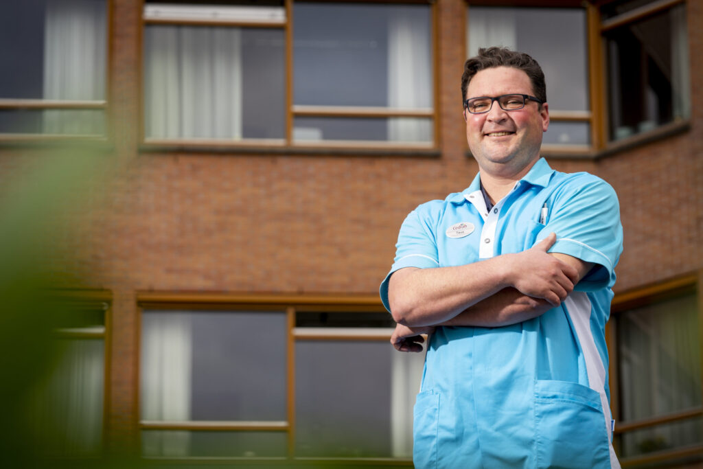 Man in blauw uniform staat met gekruiste armen voor een bakstenen gebouw met ramen. Hij glimlacht en kijkt naar de camera.