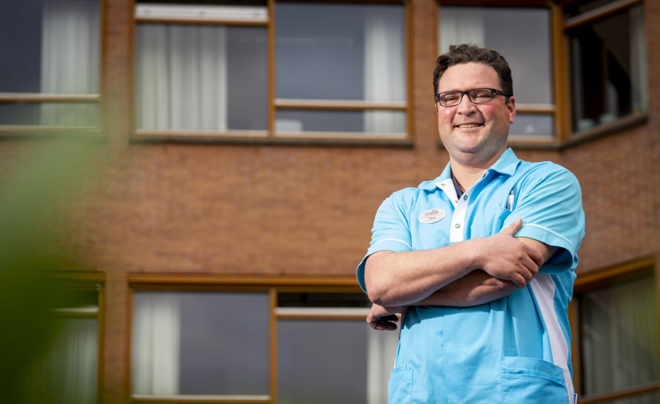 Man in blauw uniform staat met gekruiste armen voor een bakstenen gebouw met ramen. Hij glimlacht en kijkt naar de camera.
