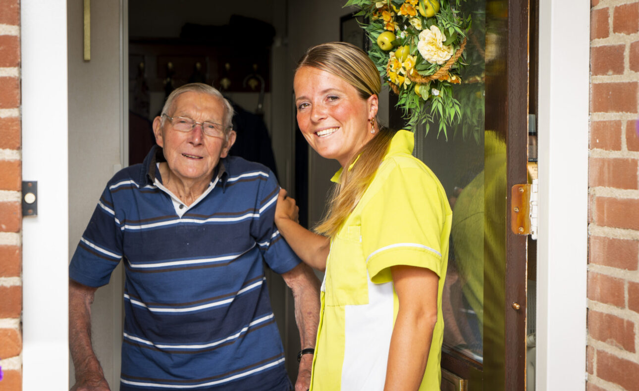 Een oudere man in een blauw gestreept shirt staat in de deuropening naast een lachende zorgverlener in een geel uniform.