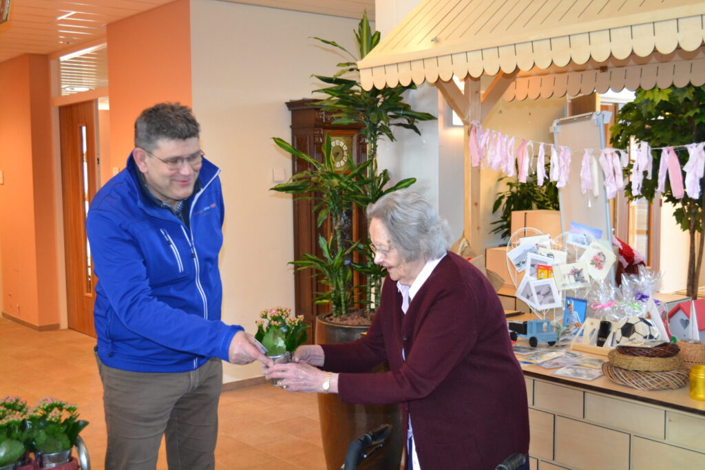 Een man in een blauwe jas overhandigt iets aan een oudere vrouw in een zorginstelling, omringd door planten en decoraties.