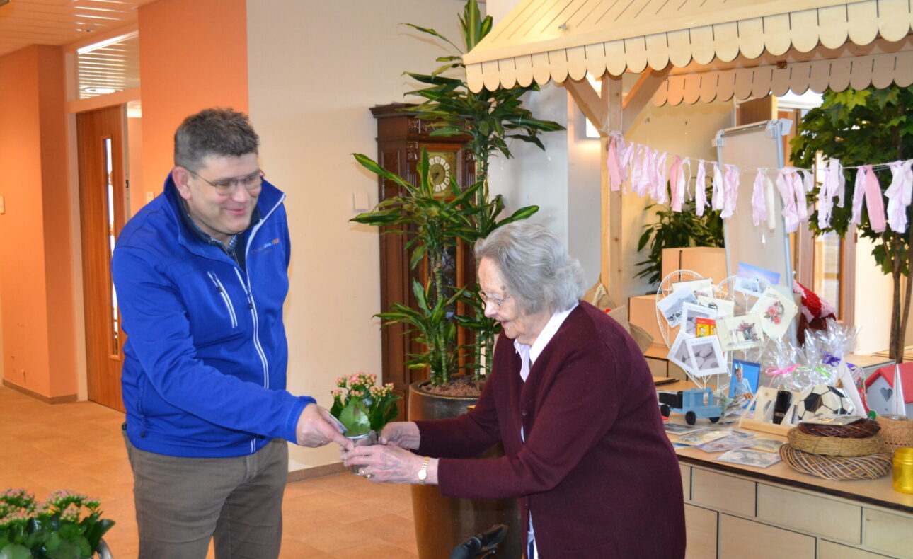 Een man in een blauwe jas overhandigt iets aan een oudere vrouw in een zorginstelling, omringd door planten en decoraties.