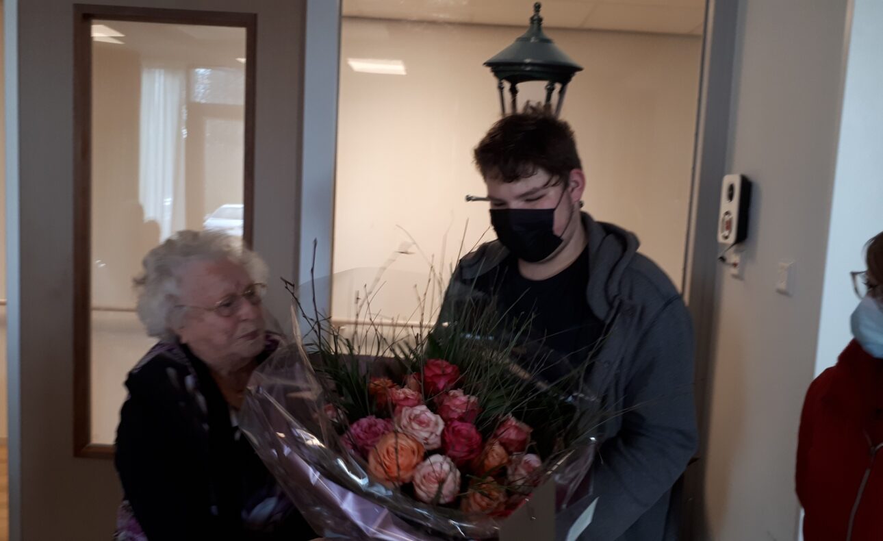 Een oudere vrouw ontvangt een grote bos rode rozen van een jongere man. Beiden dragen een mondkapje in een binnenruimte.