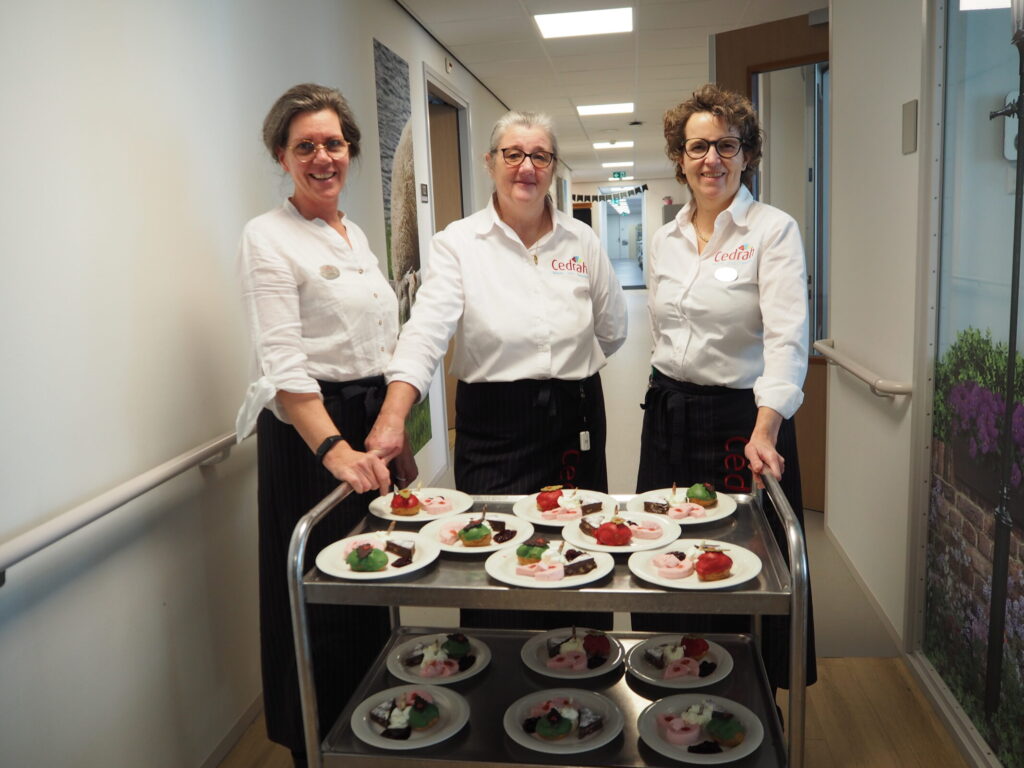 Drie vrouwen in witte blouses staan naast een serveerwagen met borden vol kleurrijke desserts in een lichte gang.