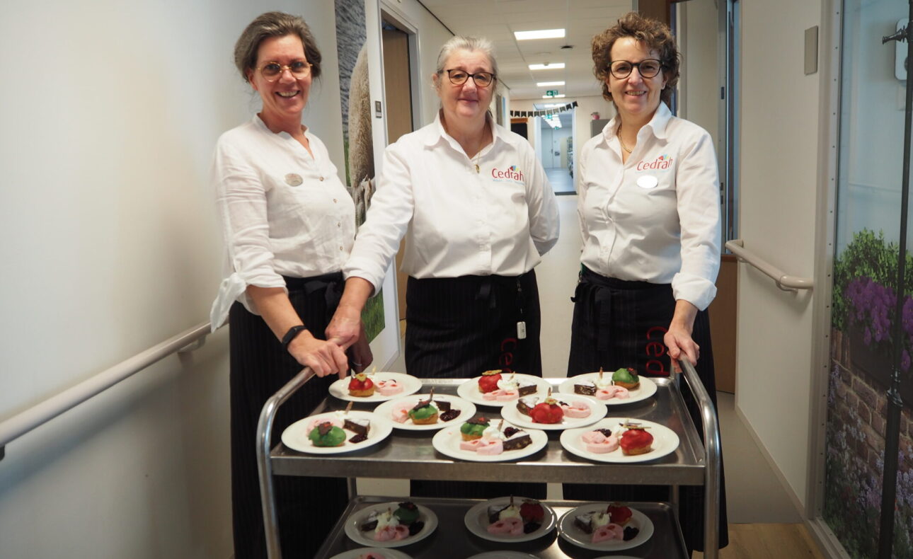 Drie vrouwen in witte blouses staan naast een serveerwagen met borden vol kleurrijke desserts in een lichte gang.