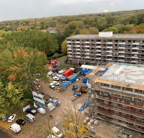 Luchtfoto van een bouwplaats met meerdere gebouwen, omringd door bomen in herfstkleuren en geparkeerde voertuigen.