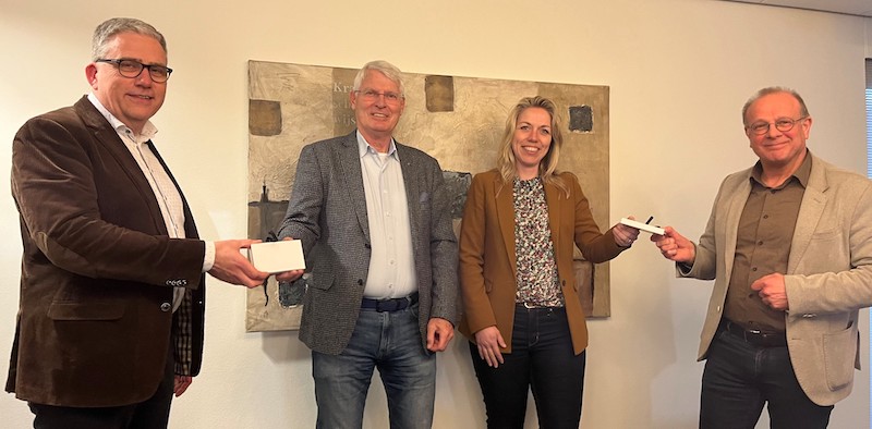Vier mensen poseren voor een schilderij aan de muur. Twee van hen houden een wit object vast en glimlachen naar de camera.
