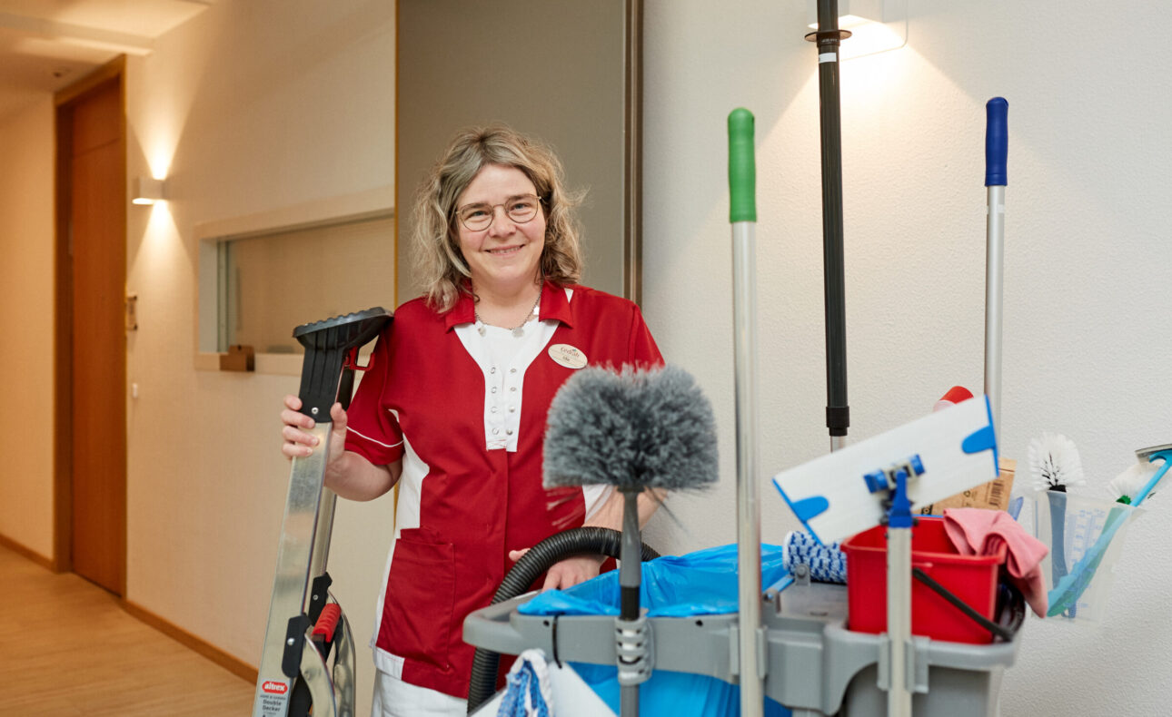 Vrouw in rood-wit uniform staat naast schoonmaakkar met diverse schoonmaakspullen in een gang van een gebouw.