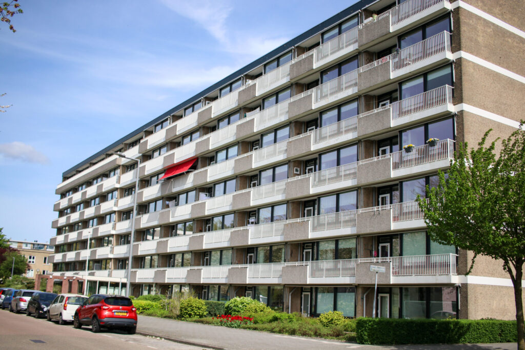 Afbeelding van een flatgebouw met meerdere verdiepingen en balkons. Er staan auto's geparkeerd langs de straat ervoor.