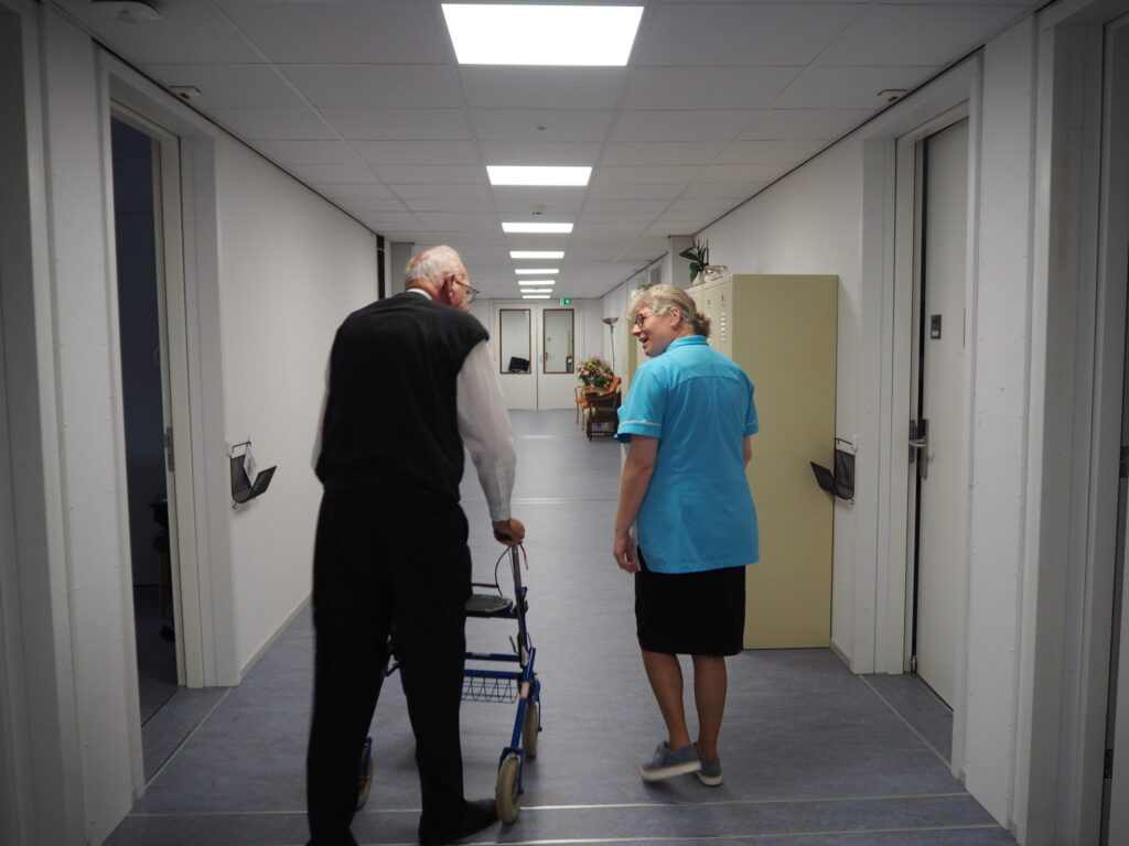 Een oudere man met een rollator loopt door een gang naast een vrouw in een blauw uniform. Ze praten met elkaar.