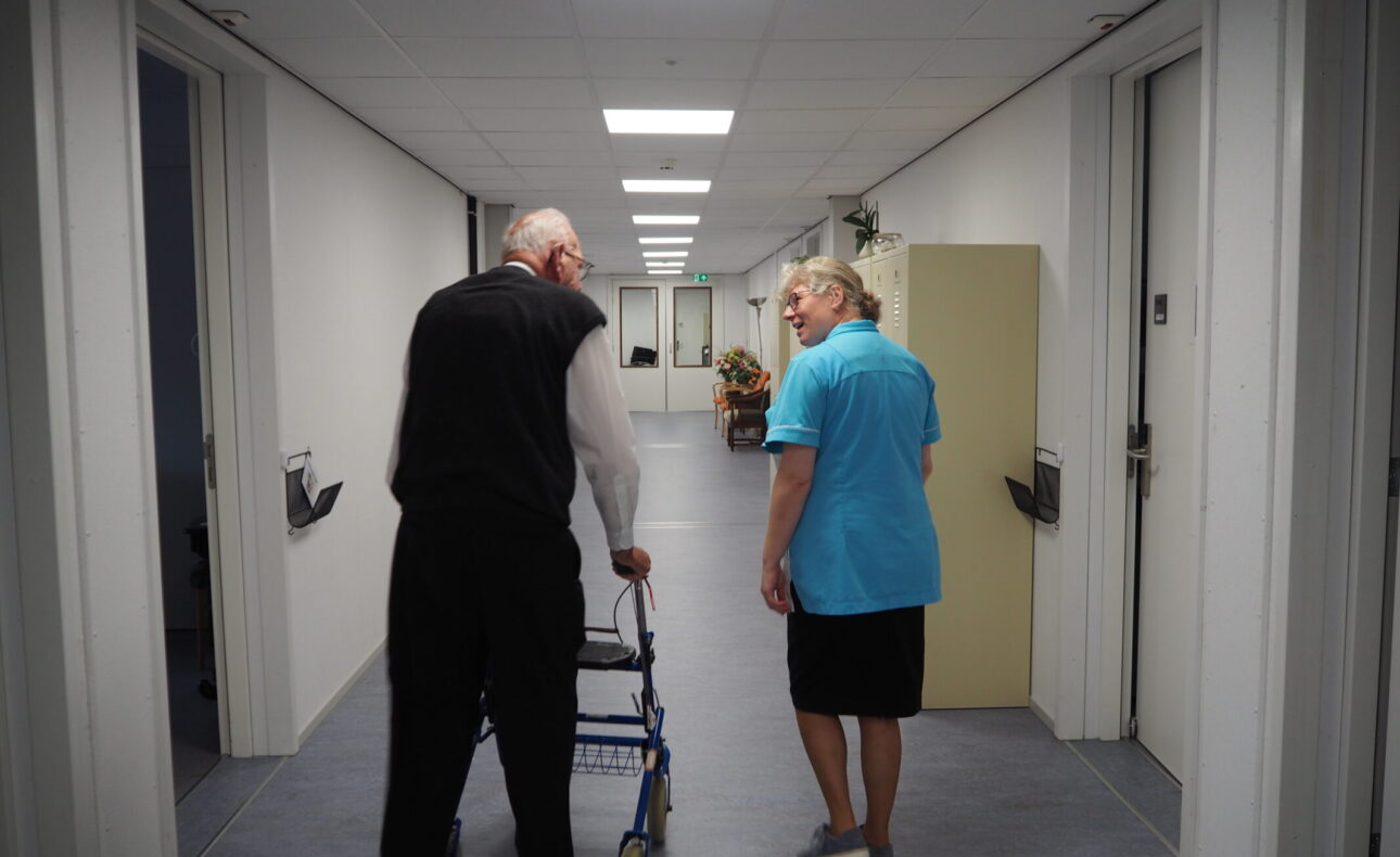 Een oudere man met een rollator loopt door een gang naast een vrouw in een blauw uniform. Ze praten met elkaar.