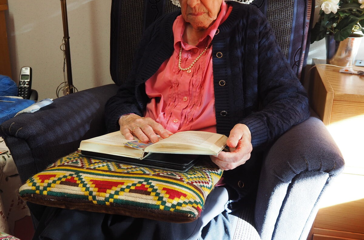 Oudere vrouw met bril leest een boek in een comfortabele stoel, omringd door kleurrijke kussens en een lamp op de achtergrond.