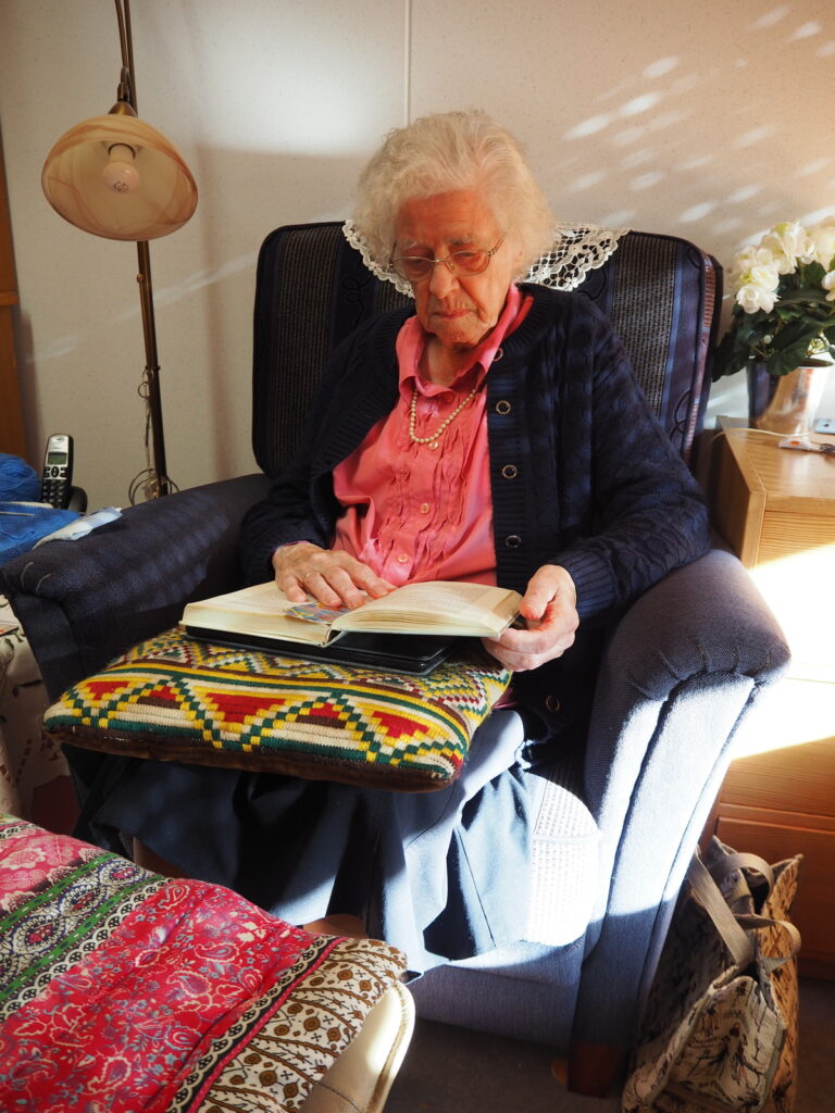 Oudere vrouw met bril leest een boek in een comfortabele stoel, omringd door kleurrijke kussens en een lamp op de achtergrond.