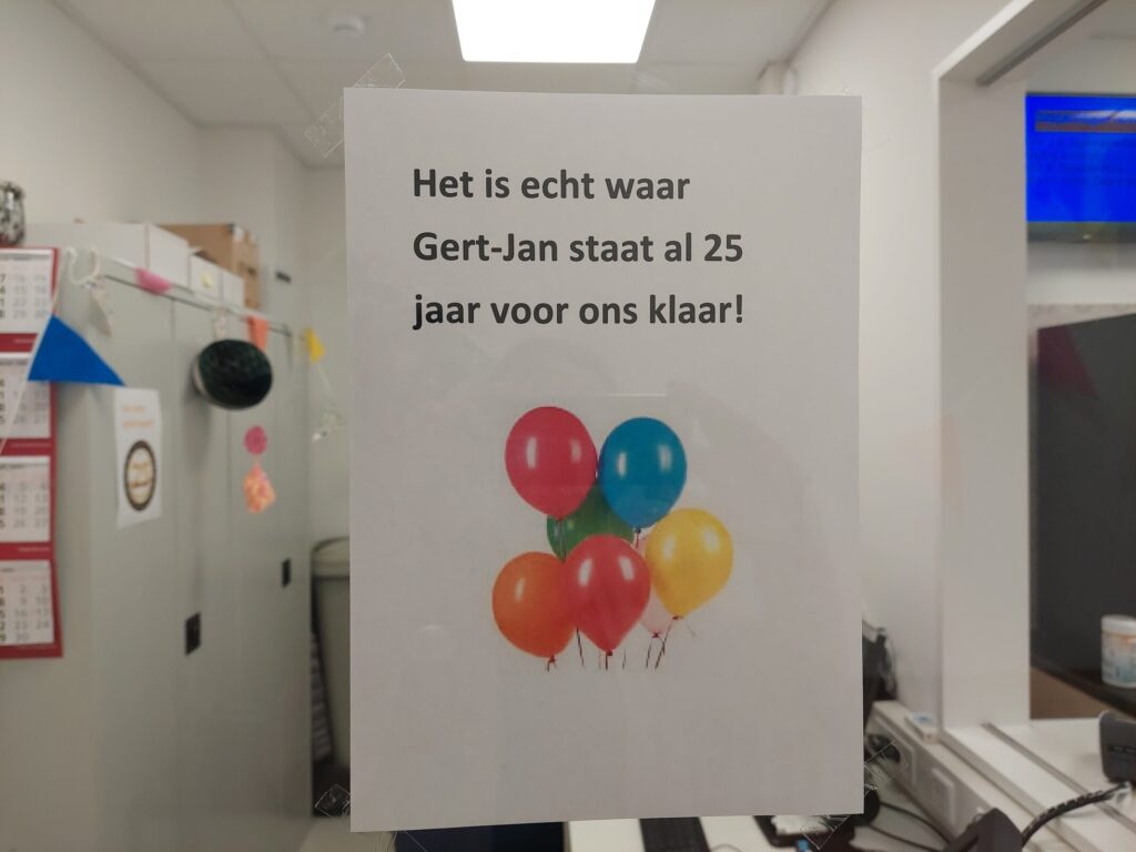 Een bord met de tekst: "Het is echt waar, Gert-Jan staat al 25 jaar voor ons klaar!" met kleurrijke ballonnen eronder.
