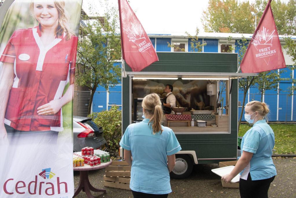 Twee zorgmedewerkers staan bij een mobiele koffiebar met Cedrah-vlaggen. Op de achtergrond is een gebouw zichtbaar.