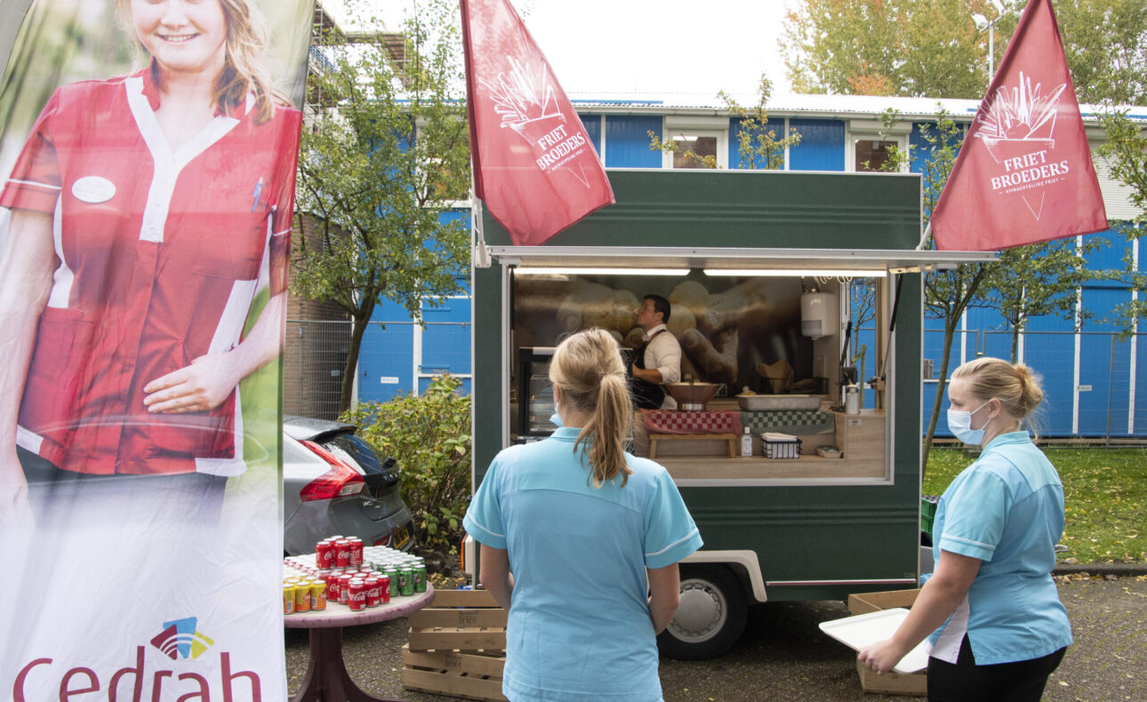 Twee zorgmedewerkers staan bij een mobiele koffiebar met Cedrah-vlaggen. Op de achtergrond is een gebouw zichtbaar.