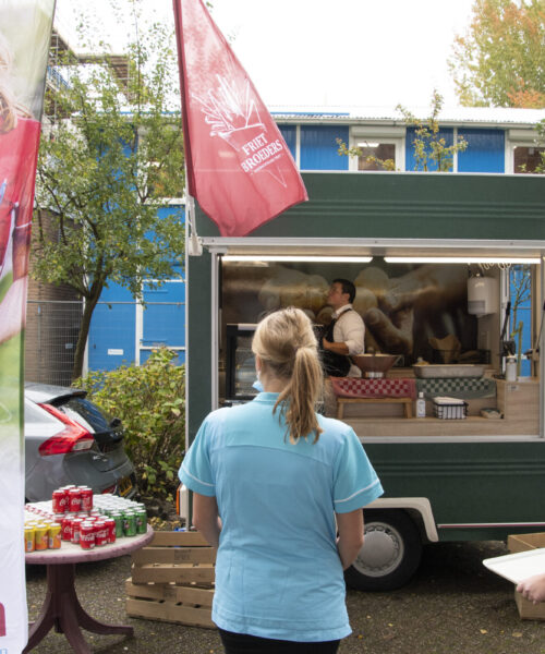 Twee zorgmedewerkers staan bij een mobiele koffiebar met Cedrah-vlaggen. Op de achtergrond is een gebouw zichtbaar.