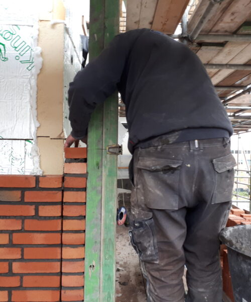 Bouwvakker met donkere kleding metselt rode bakstenen muur op steiger, omringd door bouwmaterialen en steigerplanken.