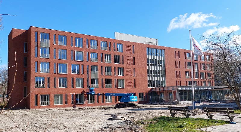Rood bakstenen gebouw met meerdere verdiepingen en grote ramen, omgeven door een bouwplaats met een blauwe kraan.
