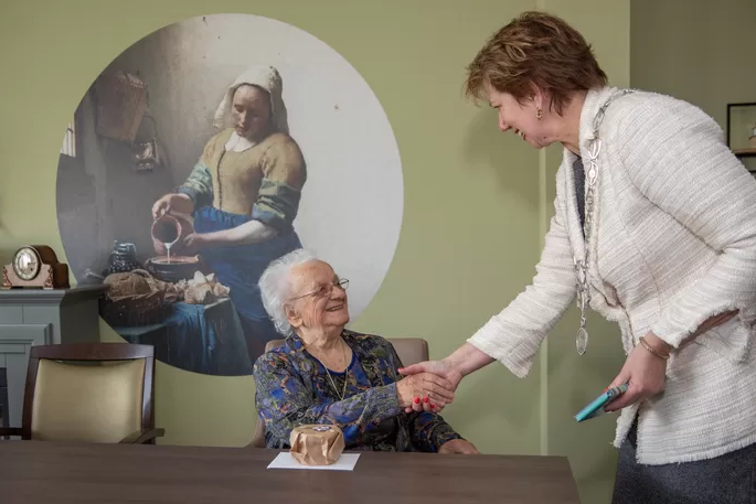 Twee vrouwen schudden handen in een kamer met een schilderij van een melkmeisje op de muur. Een vrouw zit, de ander staat.