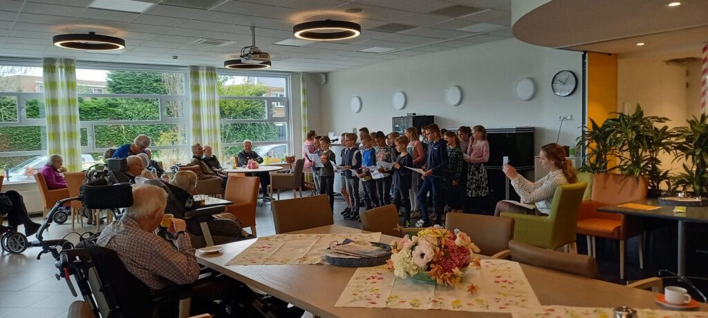 Groep kinderen zingt voor een groep ouderen in een gemeenschappelijke ruimte met tafels, stoelen en grote ramen.