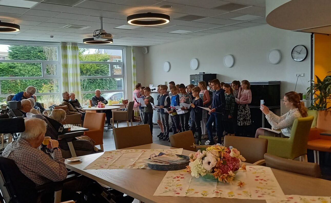 Groep kinderen zingt voor een groep ouderen in een gemeenschappelijke ruimte met tafels, stoelen en grote ramen.