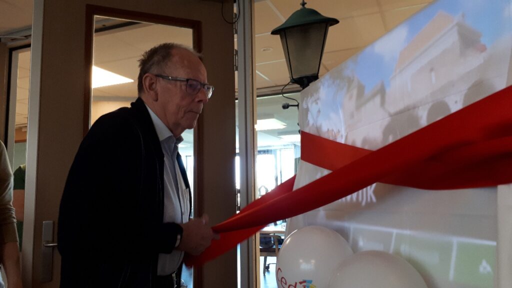 Een oudere man knipt een rood lint door tijdens een ceremonie in een gebouw, met ballonnen en een lantaarn op de achtergrond.