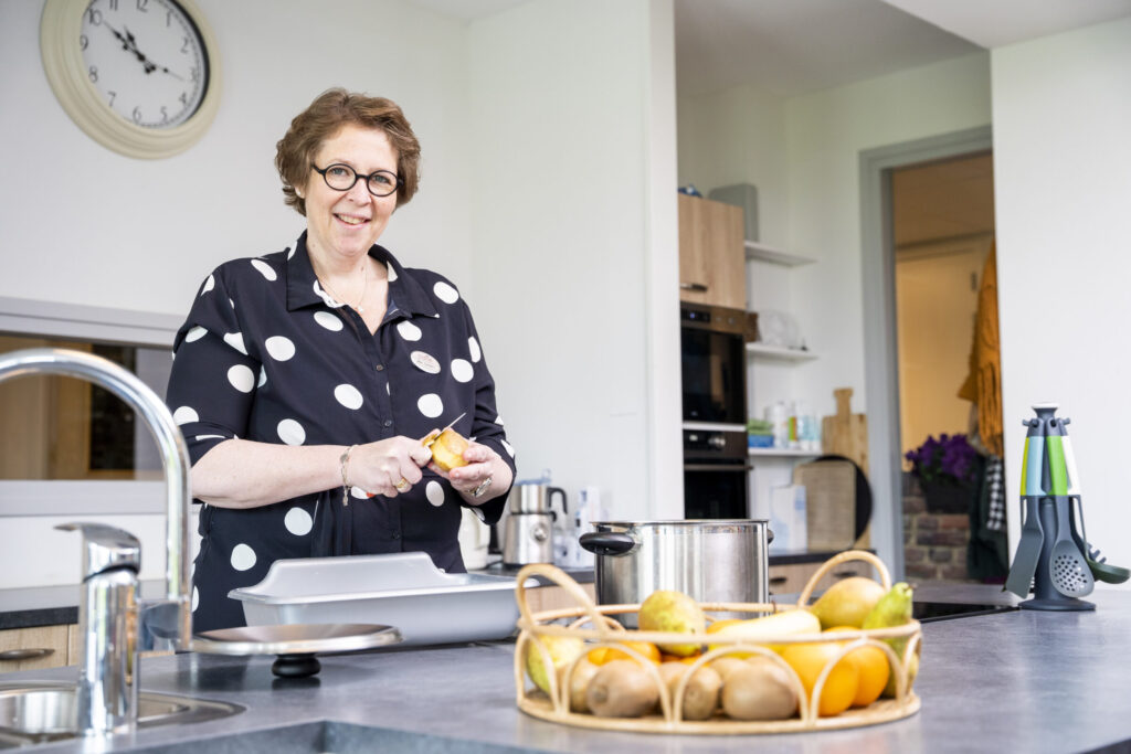 Vrouw met bril en stippenblouse schilt een sinaasappel in een moderne keuken. Op de voorgrond ligt een fruitschaal.
