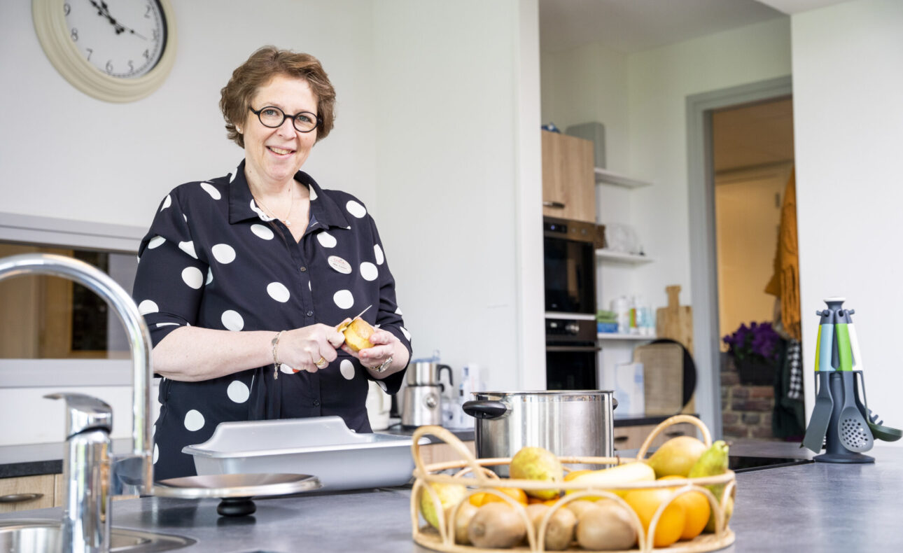 Vrouw met bril en stippenblouse schilt een sinaasappel in een moderne keuken. Op de voorgrond ligt een fruitschaal.