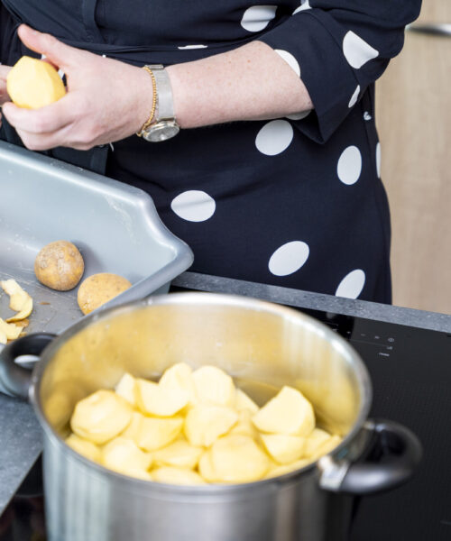 Vrouw in een donkerblauwe jurk met stippen schilt aardappelen in de keuken. Gesneden aardappelen liggen in een pan.