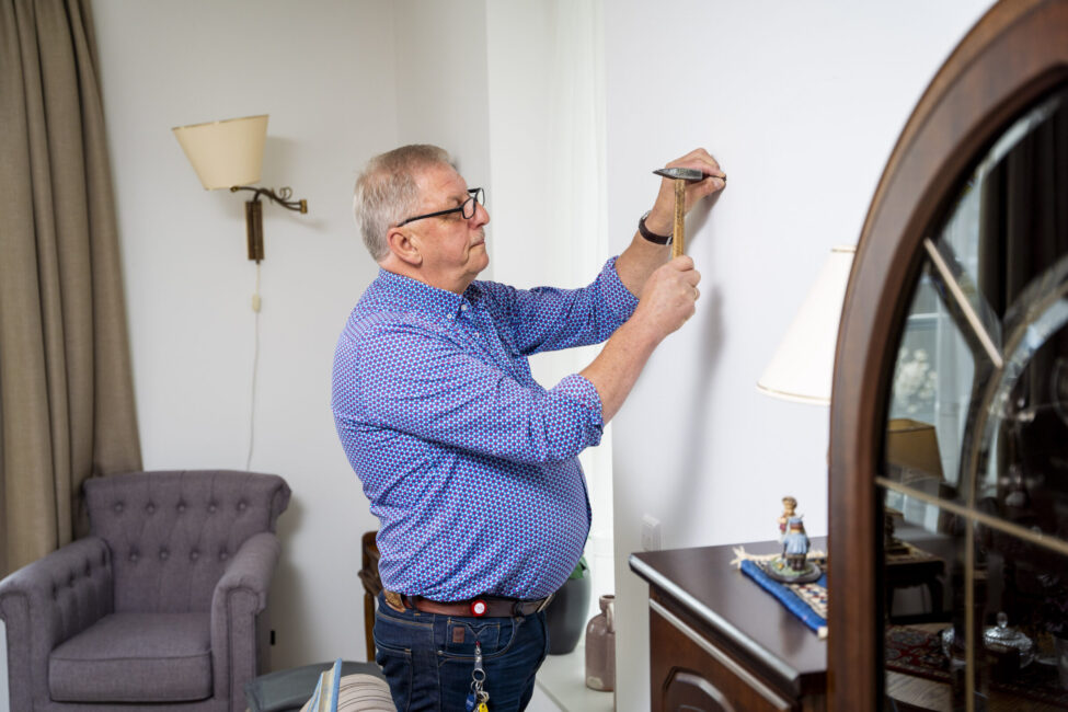 Man in blauwe blouse hangt een schilderij op aan een witte muur in een woonkamer met een grijze stoel en houten kast.
