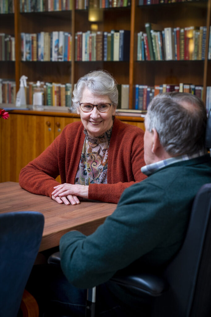 Een oudere vrouw en man zitten aan een tafel in een bibliotheek, pratend en glimlachend naar elkaar. Boeken staan op de achtergrond.