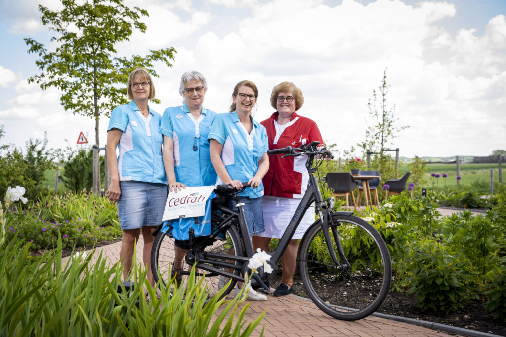 Vier vrouwen in blauwe en rode uniformen staan buiten op een pad met een fiets, omringd door groene planten en bomen.
