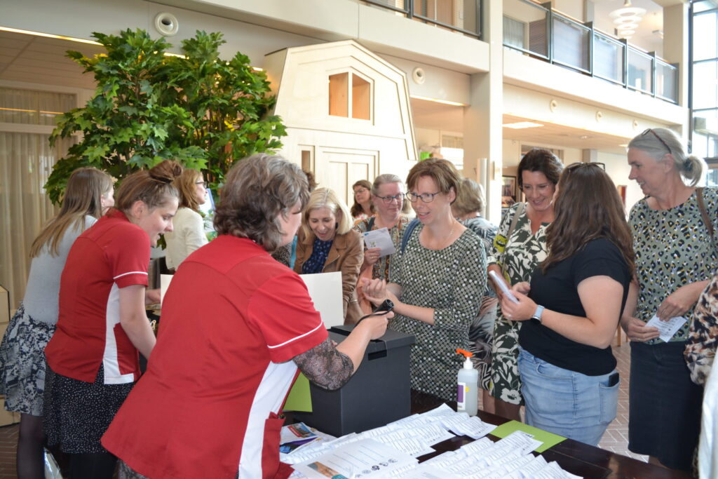 Groep mensen verzameld rond een tafel in een lichte ruimte. Een vrouw in rood uniform helpt hen met papieren en formulieren.