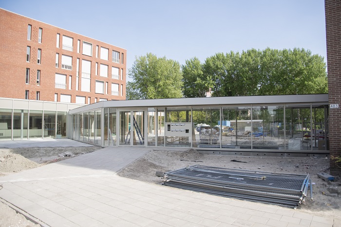Nieuwbouwproject met moderne glazen gevels en bakstenen gebouw op de achtergrond, omgeven door bomen en een bouwplaats.