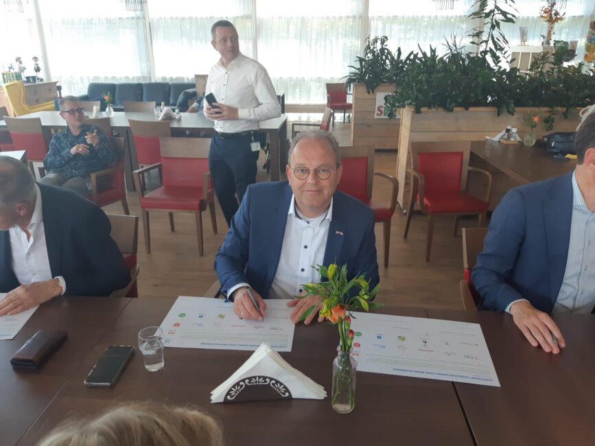 Een man in een pak ondertekent een document aan een tafel in een vergaderruimte, omringd door andere mensen en planten.