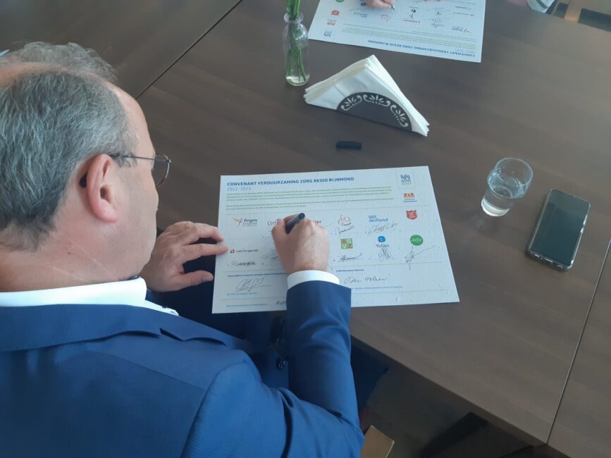 Man in blauw pak ondertekent document aan houten tafel, met een glas water en een telefoon naast hem.