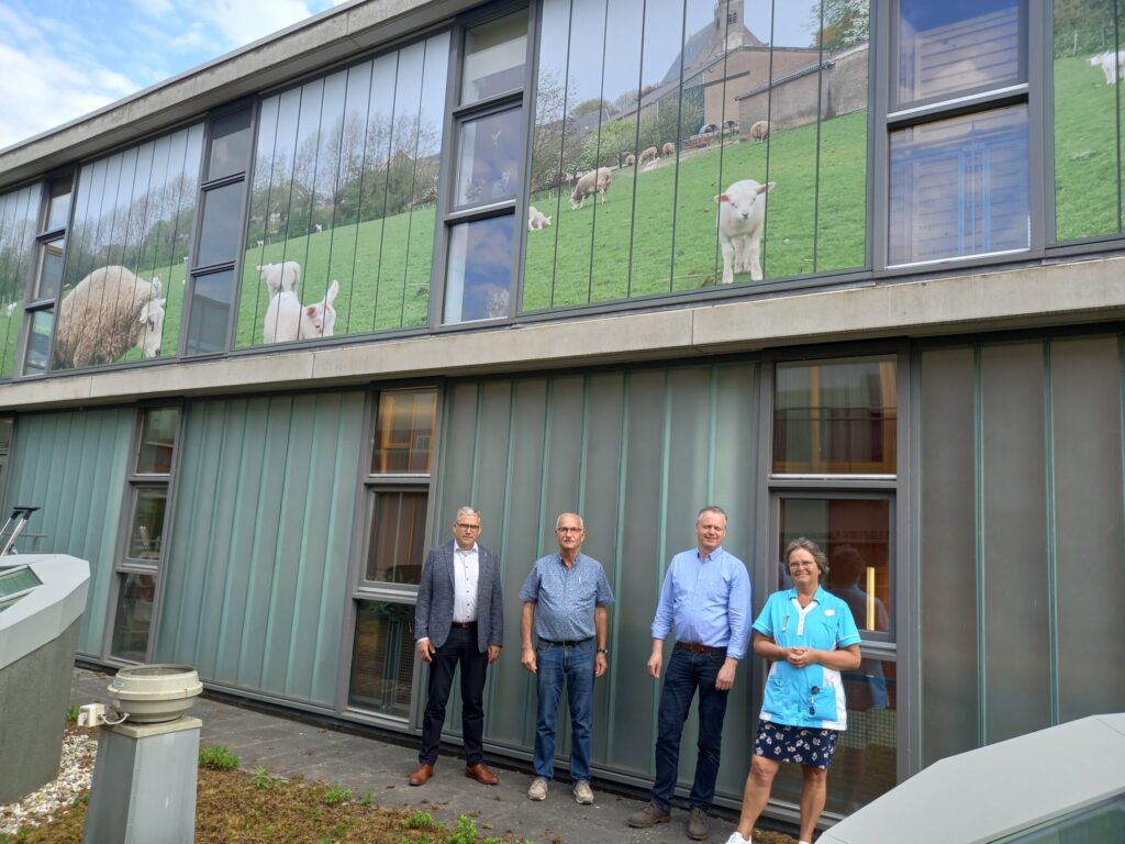 Vier mensen staan voor een gebouw met grote ramen waarop een afbeelding van een groene weide met schapen is te zien.