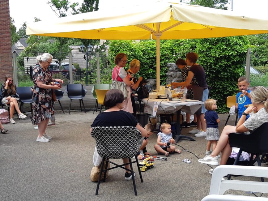 Groep mensen onder een gele parasol op een terras. Kinderen spelen op de grond, terwijl volwassenen met elkaar praten.