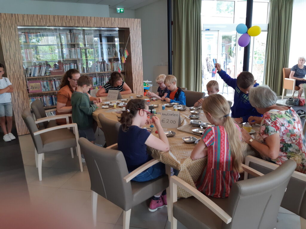 Groep kinderen en volwassenen zit rond een tafel in een gemeenschappelijke ruimte, bezig met knutselactiviteiten.