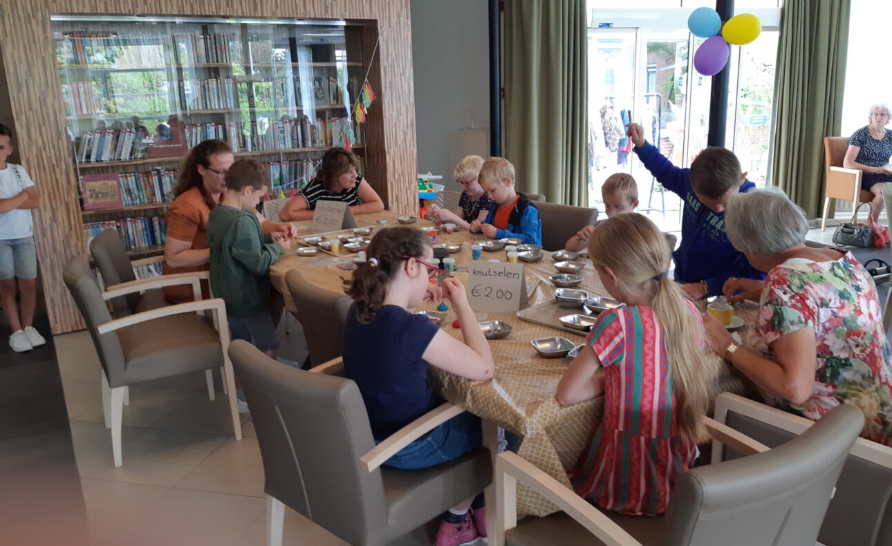 Groep kinderen en volwassenen zit rond een tafel in een gemeenschappelijke ruimte, bezig met knutselactiviteiten.