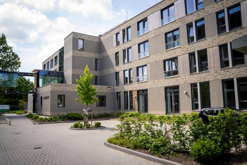 Buitenaanzicht van een modern gebouw met meerdere verdiepingen, omgeven door groen en een betegeld pad onder een blauwe lucht.